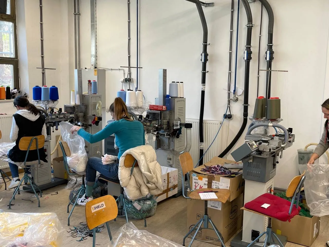 Workers seated at toe-closing (linking) machines, surrounded by cones of yarn and clear bags of material.