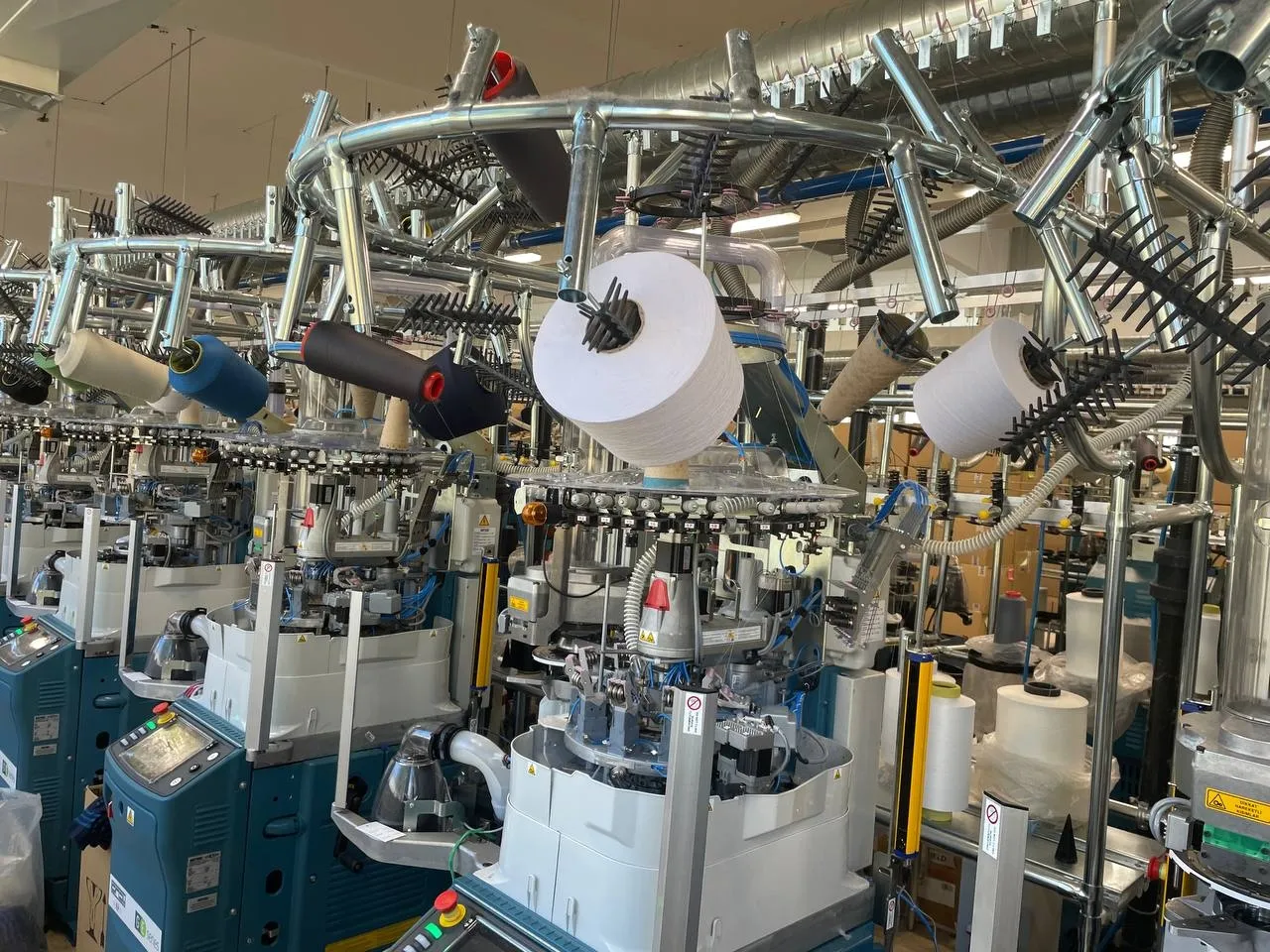 Close-up of a circular knitting machine with a large cone of white yarn threaded through the feeder, flanked by more machines down the row.